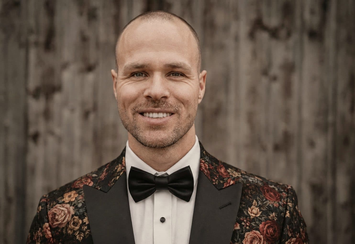 Daniel Hascher lächelt im geblümten Sakko mit Fliege vor einer Holzwand